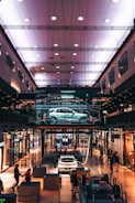 A modern shopping mall interior with an illuminated ceiling. A digital display showcases a car in the center, surrounded by various shops. People walk around and interact with exhibits, including a white car and structural components.