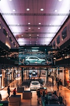 A modern shopping mall interior with an illuminated ceiling. A digital display showcases a car in the center, surrounded by various shops. People walk around and interact with exhibits, including a white car and structural components.