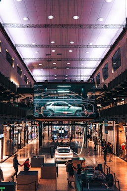 A modern shopping mall interior with an illuminated ceiling. A digital display showcases a car in the center, surrounded by various shops. People walk around and interact with exhibits, including a white car and structural components.