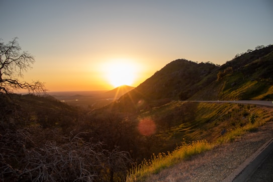 The image depicts a serene landscape at sunset with the sun low on the horizon casting a golden hue across the sky. Rolling hills are visible with verdant greenery, and a winding road runs through the area. Bare trees are silhouetted against the glowing sky, and the overall atmosphere suggests tranquility and natural beauty.