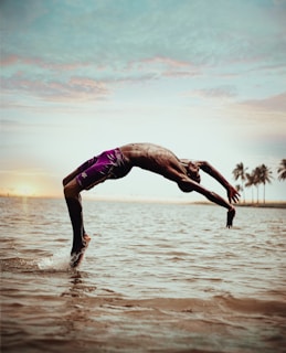 A person is performing a backflip over the water at the beach, with their body curved gracefully. The scene includes a calm ocean and a distant view of palm trees under a vibrant sky, suggesting a tropical setting.