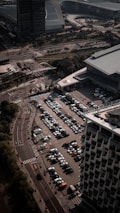 An aerial view of a large parking lot filled with rows of parked cars next to a modern building. Adjacent roads with minimal traffic and surrounding structures, including another large building, indicate an urban setting. Trees and patches of greenery are visible along the streets.