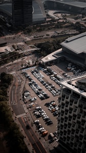 An aerial view of a large parking lot filled with rows of parked cars next to a modern building. Adjacent roads with minimal traffic and surrounding structures, including another large building, indicate an urban setting. Trees and patches of greenery are visible along the streets.