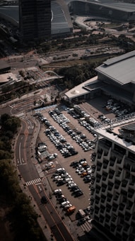 An aerial view of a large parking lot filled with rows of parked cars next to a modern building. Adjacent roads with minimal traffic and surrounding structures, including another large building, indicate an urban setting. Trees and patches of greenery are visible along the streets.