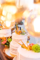 A tiered white cake is adorned with delicate patterns and topped with vibrant floral decorations including orange, yellow, and bright green elements. It sits on a wooden table, surrounded by warm lights that create a soft, atmospheric glow.