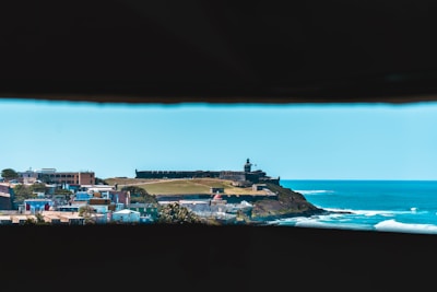 A coastal landscape featuring a historic fort on a hill, with a lighthouse atop it overlooking the ocean. In the foreground, colorful buildings form a small town, and waves are visible in the bright blue sea to the right.
