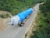 A large industrial component is being transported on a multi-axle trailer along a rural road surrounded by dense greenery. The component is covered with a bright blue tarp, indicating protection during transit. Two trucks are involved in the transport, with one leading and another following the trailer.