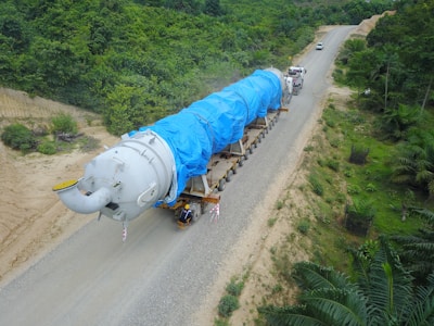A large industrial component is being transported on a multi-axle trailer along a rural road surrounded by dense greenery. The component is covered with a bright blue tarp, indicating protection during transit. Two trucks are involved in the transport, with one leading and another following the trailer.