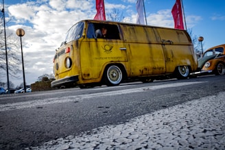 A vintage yellow van from Deutsche Bundespost is parked on a street. The body of the van appears aged with visible rust. Bright red flags are visible in the background, as well as a classic Volkswagen Beetle partially in view. Overcast sky adds to the atmosphere with street lamps and trees lining the road.