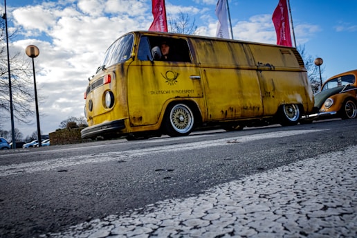 A vintage yellow van from Deutsche Bundespost is parked on a street. The body of the van appears aged with visible rust. Bright red flags are visible in the background, as well as a classic Volkswagen Beetle partially in view. Overcast sky adds to the atmosphere with street lamps and trees lining the road.