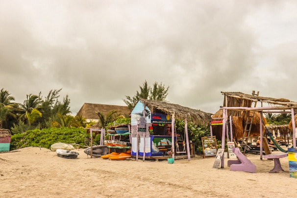 A rustic beachfront setup features a small hut with a thatched roof adorned with colorful signs, surrounded by paddleboards and beach equipment. Palm trees and dense greenery form a backdrop, while the sky is overcast with heavy clouds, suggesting an impending storm. Relaxation benches painted in vibrant colors add a touch of charm to the scene.