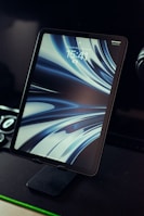 A tablet is displayed on a stand with a digital art wallpaper featuring fluid, abstract blue and white swirls. The screen shows the time 15:41 and a lock icon in the center, indicating it's in the lock screen mode. The device is in a dark environment with some green accent lighting seen at the bottom of the image.