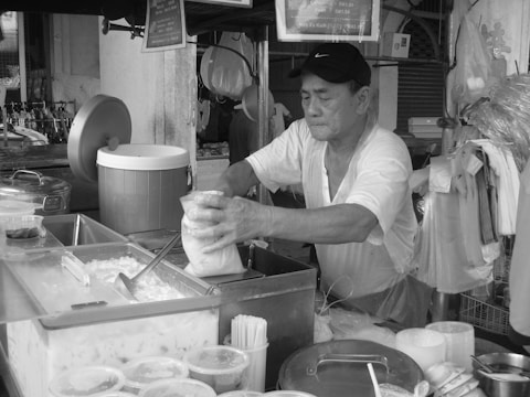 An elderly man wearing a cap works at a street food stall, handling a plastic bag filled with food. The stall is cluttered with containers, coolers, and plastic cups. Behind him, various tools and utensils hang on the wall.