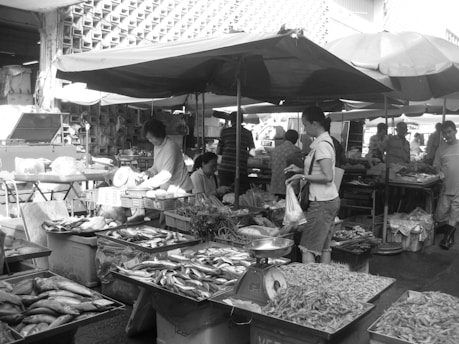 A bustling outdoor market scene with vendors selling a variety of fresh fish and seafood. Multiple tables display different types of fish and shrimp, with customers interacting with the sellers. Umbrellas provide shade as people go about their shopping. Stalls are busy with activity and people examining the produce.