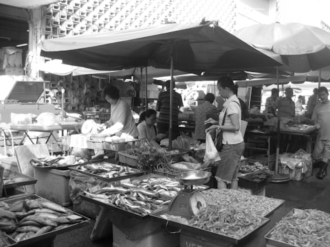 A bustling outdoor market scene with vendors selling a variety of fresh fish and seafood. Multiple tables display different types of fish and shrimp, with customers interacting with the sellers. Umbrellas provide shade as people go about their shopping. Stalls are busy with activity and people examining the produce.