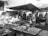 A bustling outdoor market scene with vendors selling a variety of fresh fish and seafood. Multiple tables display different types of fish and shrimp, with customers interacting with the sellers. Umbrellas provide shade as people go about their shopping. Stalls are busy with activity and people examining the produce.