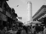 A bustling outdoor market scene with numerous people walking between stalls. The crowd is set against a backdrop of urban buildings, including a tall skyscraper. Stalls have umbrellas and a variety of goods displayed, indicating a lively marketplace atmosphere.