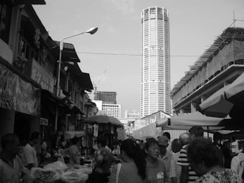 A bustling outdoor market scene with numerous people walking between stalls. The crowd is set against a backdrop of urban buildings, including a tall skyscraper. Stalls have umbrellas and a variety of goods displayed, indicating a lively marketplace atmosphere.