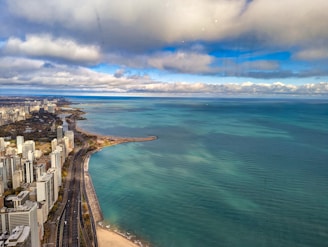 A coastal city skyline with high-rise buildings along a curving shoreline. The expansive body of water is a shade of blue-green, contrasting with the partly cloudy sky. The highway runs parallel to the coast, with visible infrastructure and urban development.
