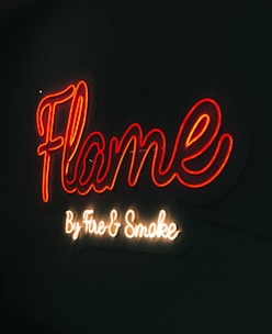 A neon sign with the word 'Flame' written in vibrant red cursive script. Below it, in a smaller white cursive font, the words 'By Fire & Smoke' are displayed. The sign is illuminated against a dark background, creating a vivid contrast.