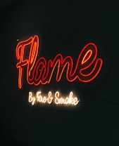 A neon sign with the word 'Flame' written in vibrant red cursive script. Below it, in a smaller white cursive font, the words 'By Fire & Smoke' are displayed. The sign is illuminated against a dark background, creating a vivid contrast.