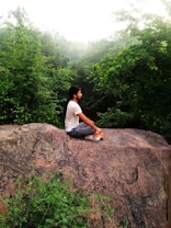 A person is sitting cross-legged on a large rock, surrounded by lush green trees. The setting appears calm and serene, with the misty background adding an ethereal quality to the scene.