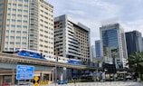 A cityscape featuring a modern light rail transit train traveling on an elevated track. The background includes several tall buildings, including Menara Cosway with reflective glass windows. A signpost with directions to local landmarks such as Petaling Jaya can be seen, along with palm trees and other urban infrastructure.