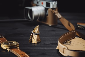 A luxurious scene featuring a gold spiral-shaped object adorned with a dark ring, set on a dark wooden surface. Nearby, a vintage camera with brown leather accents and a classic wristwatch with a brown leather strap are elegantly arranged. A leather strap frames an old letter with intricate script, adding to the nostalgic atmosphere.
