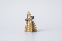 A gold, pyramid-like object with a textured surface stands upright. It has a black or dark gray, intricately crafted ring placed around it. The background is a simple, clean white, emphasizing the object's elegance.