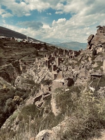 A picturesque scene of ancient ruins on a hillside, with stone structures in various states of decay spread across the steep, lush landscape. The backdrop features rolling mountains and a partly cloudy sky, creating a dramatic and historical atmosphere.
