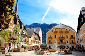 A picturesque European village square featuring colorful buildings with charming balconies and vibrant vegetation. People are leisurely sitting at outdoor caf&eacute; seating, and a bicycle is parked nearby. A mountain range with clear blue skies serves as a stunning backdrop.