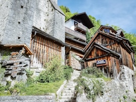 An old, rustic structure with wooden and stone elements is nestled against a hillside. The architecture includes a mix of stone walls and wooden facades, with stacked firewood visible. Lush greenery and flowering plants surround the steps leading up the hill. Signs with words such as 'Gaiswandweg' and 'Zur Kirche' are displayed prominently.