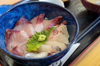 A vibrant dish featuring slices of raw fish, laid meticulously on rice in a deep, blue ceramic bowl. The fish appears fresh with hues of pink and red, and is adorned with a piece of green shiso leaf and a dollop of wasabi in the center for garnish.