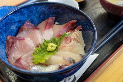 A vibrant dish featuring slices of raw fish, laid meticulously on rice in a deep, blue ceramic bowl. The fish appears fresh with hues of pink and red, and is adorned with a piece of green shiso leaf and a dollop of wasabi in the center for garnish.
