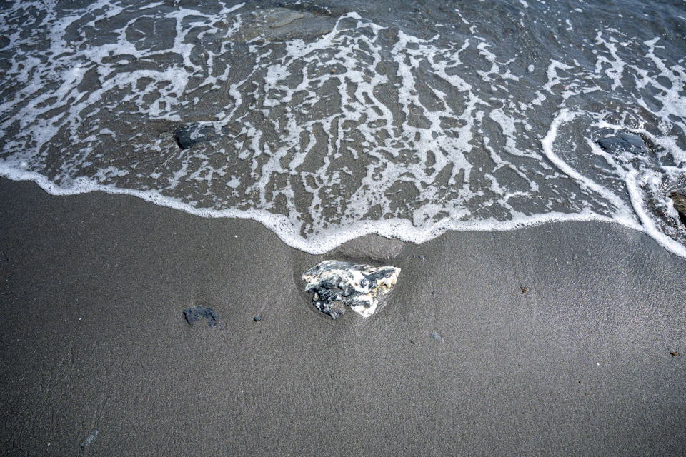 The image depicts a serene beach scene with light beige sand and dark gray water. The waves are crashing onto the shore, creating a frothy white foam that contrasts with the darker water. A large rock is partially submerged under the water's surface, adding an element of ruggedness to the otherwise smooth landscape.

The perspective of the image is from above, providing a bird's eye view of the beach and its features. The sky forms part of the background, subtly visible through the top left corner of the photo.