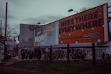 A street scene with large billboards, one promoting 102.7 NOW! radio and another advertising 'Here, There, Everywhere' by Pattison. The area features graffiti on walls and a sign for a T&Q laundromat on a pole. The environment appears urban and slightly rundown with sparse vegetation.