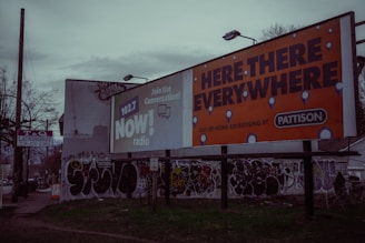 A street scene with large billboards, one promoting 102.7 NOW! radio and another advertising 'Here, There, Everywhere' by Pattison. The area features graffiti on walls and a sign for a T&Q laundromat on a pole. The environment appears urban and slightly rundown with sparse vegetation.
