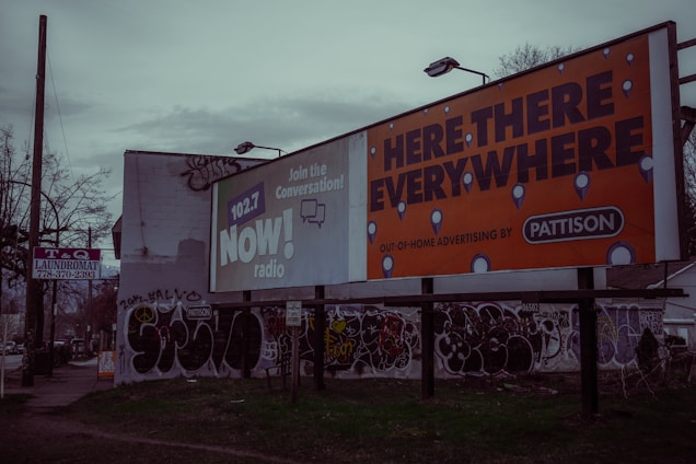 A street scene with large billboards, one promoting 102.7 NOW! radio and another advertising 'Here, There, Everywhere' by Pattison. The area features graffiti on walls and a sign for a T&Q laundromat on a pole. The environment appears urban and slightly rundown with sparse vegetation.