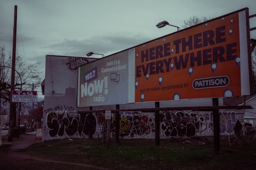 A street scene with large billboards, one promoting 102.7 NOW! radio and another advertising 'Here, There, Everywhere' by Pattison. The area features graffiti on walls and a sign for a T&Q laundromat on a pole. The environment appears urban and slightly rundown with sparse vegetation.