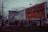 A street scene with large billboards, one promoting 102.7 NOW! radio and another advertising 'Here, There, Everywhere' by Pattison. The area features graffiti on walls and a sign for a T&Q laundromat on a pole. The environment appears urban and slightly rundown with sparse vegetation.