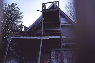 An abandoned, dilapidated house with a weathered facade and broken balcony. Graffiti is visible on the lower part of the structure. The roof and wooden elements appear damaged, and there are leafless trees surrounding the building.