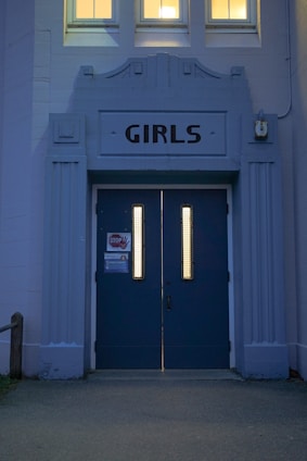 A blue double door with vertical rectangular windows, labeled 'GIRLS' above. The building facade has classical-style architecture with decorative elements in the same blue color. Two windows are visible above the label and emit a warm glow. Attached to the left door is a sign with a 'STOP' hand graphic in red.