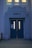 A blue double door with vertical rectangular windows, labeled 'GIRLS' above. The building facade has classical-style architecture with decorative elements in the same blue color. Two windows are visible above the label and emit a warm glow. Attached to the left door is a sign with a 'STOP' hand graphic in red.