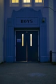 An entrance with two tall doors labeled 'BOYS' featuring vertical windows that are illuminated from the inside. The architecture is structured with an art deco style facade in bluish tones, and three small windows above cast a warm yellow light.