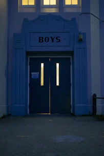 An entrance with two tall doors labeled 'BOYS' featuring vertical windows that are illuminated from the inside. The architecture is structured with an art deco style facade in bluish tones, and three small windows above cast a warm yellow light.