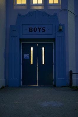 An entrance with two tall doors labeled 'BOYS' featuring vertical windows that are illuminated from the inside. The architecture is structured with an art deco style facade in bluish tones, and three small windows above cast a warm yellow light.