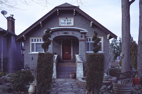 A quaint, single-story house with a steep roof and textured exterior walls. The entrance features a dark wooden door framed by two decorative evergreens. Large windows flank the doorway, and a stone pathway leads up to the steps. Surrounding the walkway, potted plants and small shrubs add greenery to the scene. The house is set in a suburban environment with trees and neighboring houses visible in the background, giving an overall rustic charm.