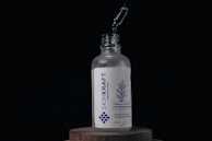 A glass bottle labeled 'SKINKRAFT LABORATORIES' stands on a wooden surface. Above the bottle, a dropper releases a droplet of liquid into the open neck. The label features a blue design of rosemary and mentions 'Rosemary with Caffeine Pro-Growth Hair Serum' in white and blue colors.