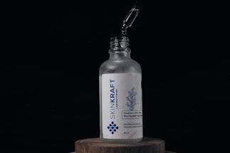 A glass bottle labeled 'SKINKRAFT LABORATORIES' stands on a wooden surface. Above the bottle, a dropper releases a droplet of liquid into the open neck. The label features a blue design of rosemary and mentions 'Rosemary with Caffeine Pro-Growth Hair Serum' in white and blue colors.