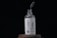 A glass bottle labeled 'SKINKRAFT LABORATORIES' stands on a wooden surface. Above the bottle, a dropper releases a droplet of liquid into the open neck. The label features a blue design of rosemary and mentions 'Rosemary with Caffeine Pro-Growth Hair Serum' in white and blue colors.